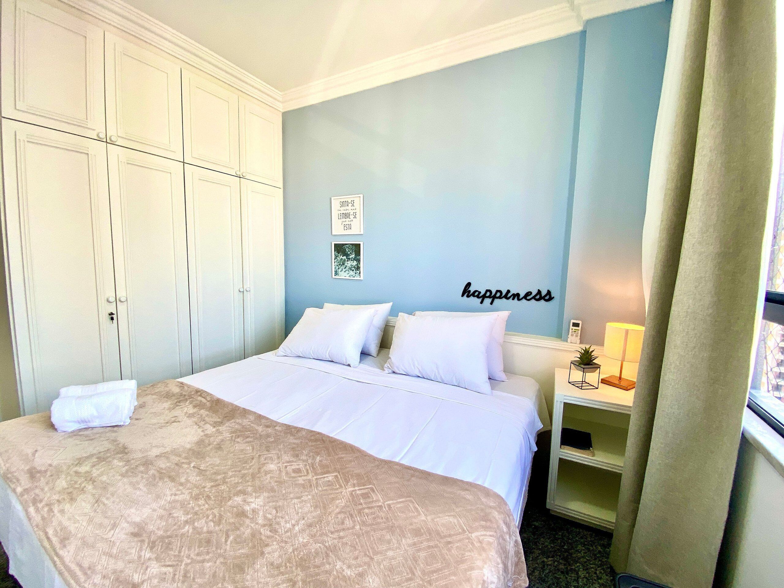 A bedroom with a bed, a wardrobe, a bedside table with a lamp, and a wall with a 'happiness' sign.