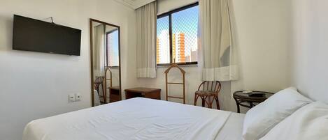 Habitación de hotel con una cama, televisión montada en la pared, ventana con cortinas, una mesita y una silla.