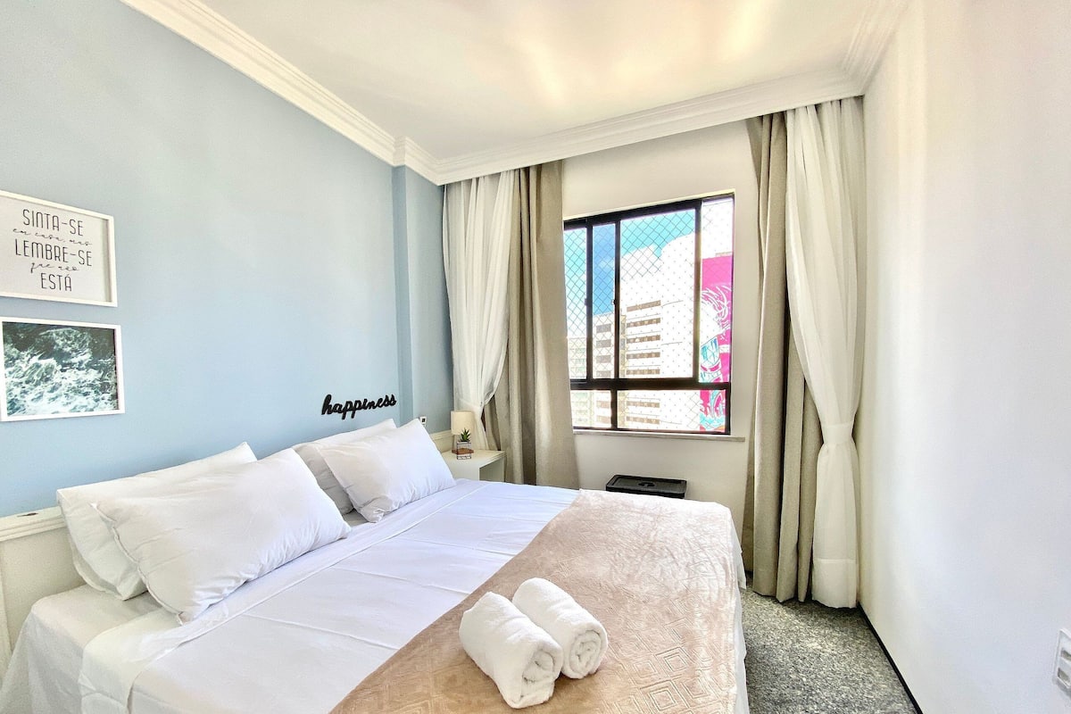 A hotel room with a bed, a window with curtains, a wall with a framed quote, and a small table with a lamp.