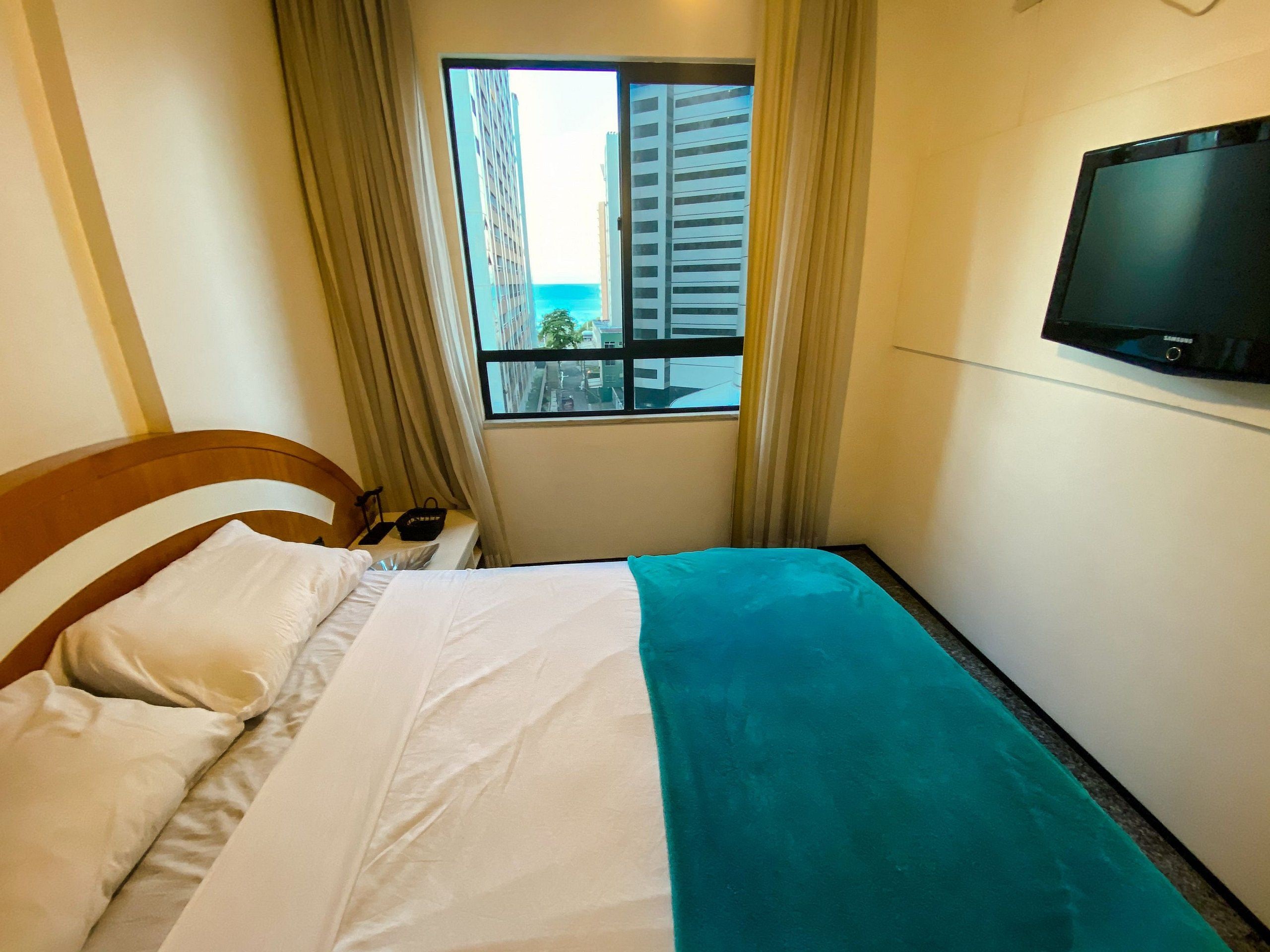 Quarto de hotel com cama, televisão, janela com cortinas e vista para edifícios.