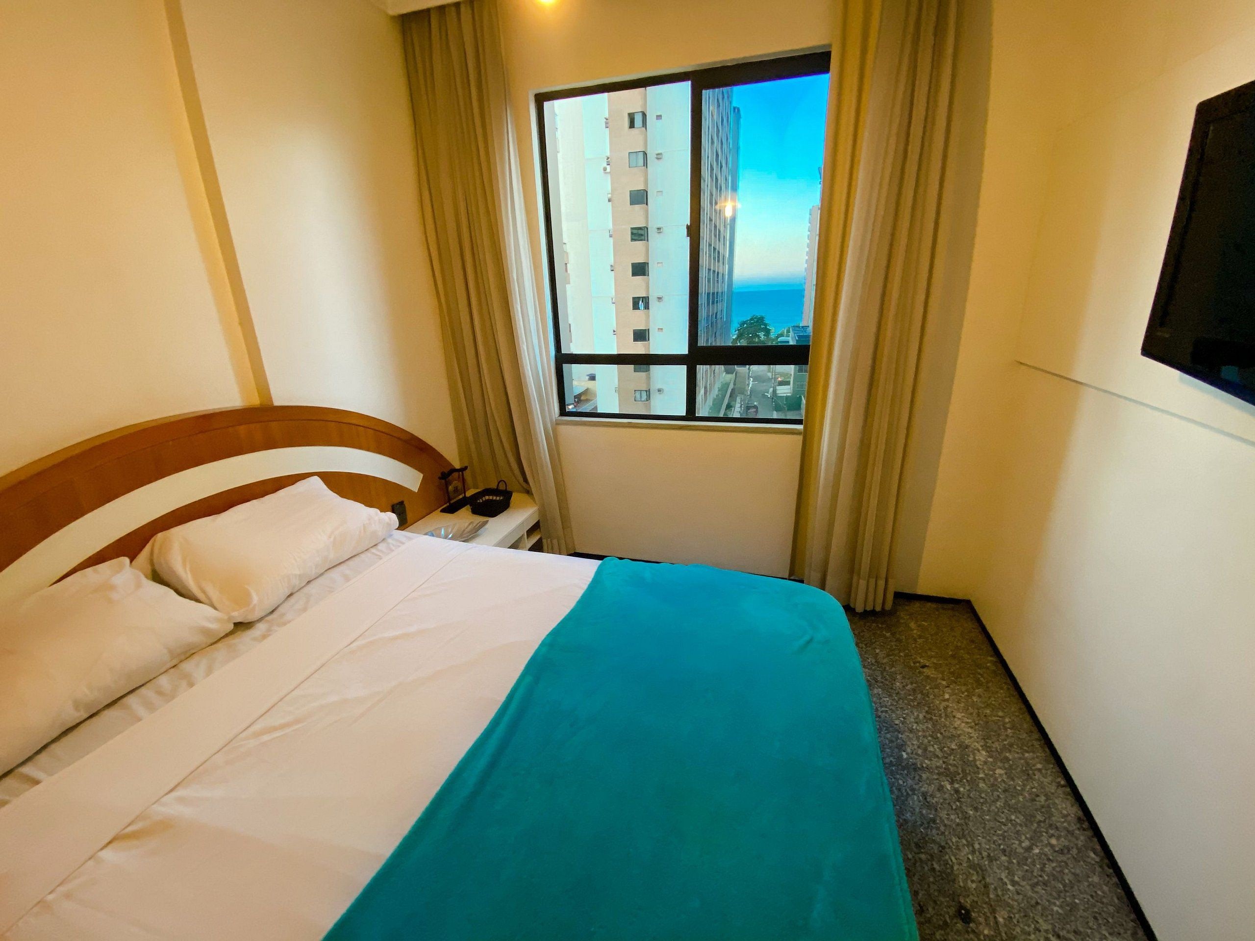 Une chambre d’hôtel avec un lit, une fenêtre avec des rideaux, une table de chevet et un téléviseur fixé au mur.