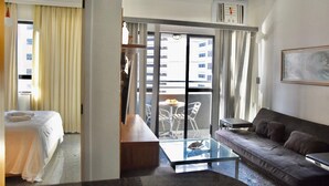 Habitación de hotel con una cama, una mesita de centro de cristal, un sofá, un balcón con mesa y sillas, y un ventanal grande con cortinas.
