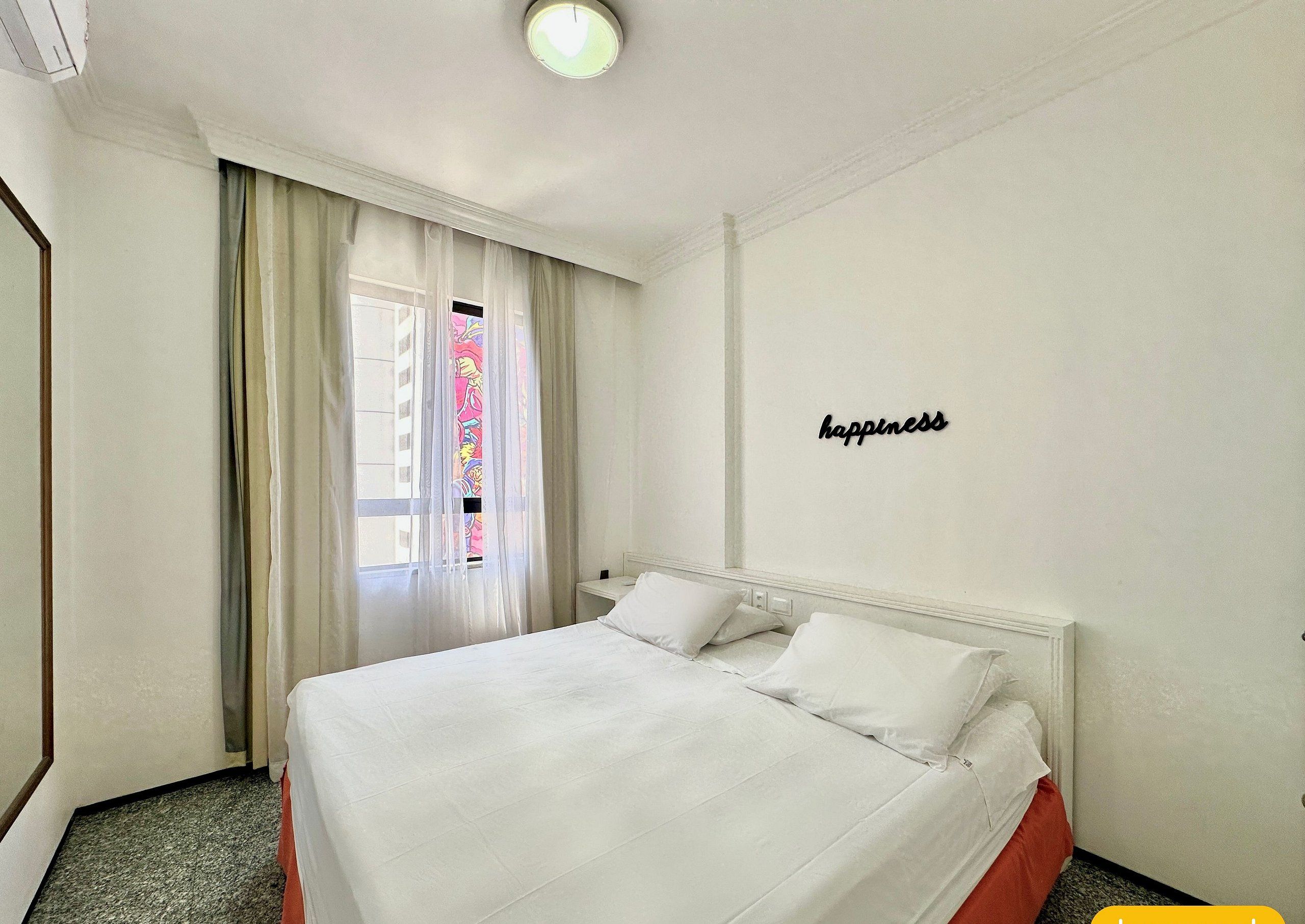 A hotel room with a bed, a window with sheer curtains, a wall-mounted light fixture, and a 'happiness' sign.