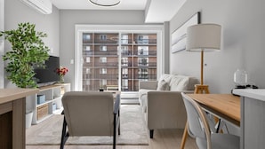 A modern apartment with a dining area, a sofa, a TV, and a large window with a view of a brick building. - 2BR | Mins to Stampede | UG Park | AC | BRAND NEW (Calgary)