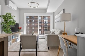 A modern apartment with a dining area, a sofa, a TV, and a large window with a view of a brick building. - 2BR | Mins to Stampede | UG Park | AC | BRAND NEW (Calgary)
