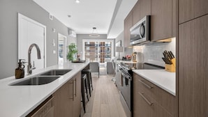 A modern kitchen with a central island, stainless steel sink, and wooden cabinets. - 2BR | Mins to Stampede | UG Park | AC | BRAND NEW (Calgary)