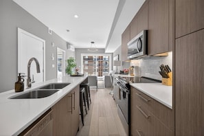 A modern kitchen with a central island, stainless steel sink, and wooden cabinets. - 2BR | Mins to Stampede | UG Park | AC | BRAND NEW (Calgary)
