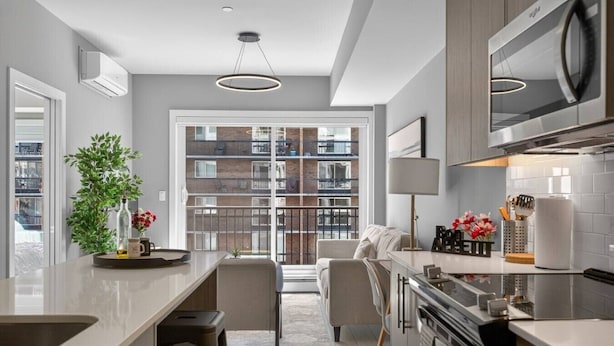 A modern kitchen with a central island, a dining area with a sofa, and a view of a balcony with a railing. - 2BR | Mins to Stampede | UG Park | AC | BRAND NEW (Calgary)