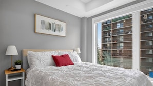 A bedroom with a large bed, a nightstand with a lamp, a framed abstract painting, and a window with a view of a building. - 2BR | Mins to Stampede | UG Park | AC | BRAND NEW (Calgary)