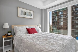 A bedroom with a large bed, a nightstand with a lamp, a framed abstract painting, and a window with a view of a building. - 2BR | Mins to Stampede | UG Park | AC | BRAND NEW (Calgary)