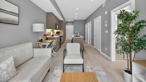 A modern living room with a sofa, a coffee table, and a dining area with a table and chairs. - 2BR | Mins to Stampede | UG Park | AC | BRAND NEW (Calgary)