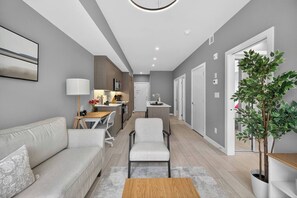 A modern living room with a sofa, a coffee table, and a dining area with a table and chairs. - 2BR | Mins to Stampede | UG Park | AC | BRAND NEW (Calgary)