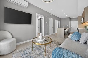 Ein modernes Wohnzimmer mit einer Couch, einem Couchtisch und einem Fernseher. Es gibt Türen zu anderen Räumen, eine Küchenzeile und ein Badezimmer.