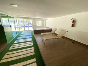 A waiting area with a green carpet, a fire extinguisher on the wall, and a view of the outside through large windows. - VG Fun Praia do Futuro - CE (Fortaleza)
