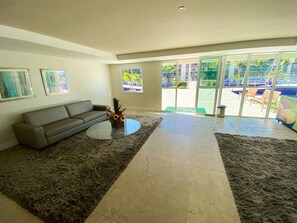 A living room with a sofa, a coffee table, a rug, and a view of an outdoor area with chairs and a pool. - VG Fun Praia do Futuro - CE (Fortaleza)