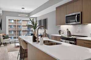 A modern kitchen with a central island, stainless steel appliances, and a view of a residential building. - Luxury 2 Bedroom | Min to Stampede | UG Prk | AC (Calgary)