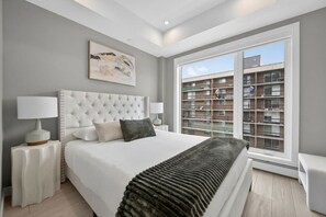 A bedroom with a large bed, two bedside tables with lamps, a window with a view of a building, and a painting on the wall. - Luxury 2 Bedroom | Min to Stampede | UG Prk | AC (Calgary)
