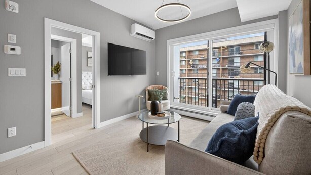 Ein modernes Wohnzimmer mit Sofa, Couchtisch und Fernseher. Ein Balkon bietet Blick auf umliegende Gebäude.