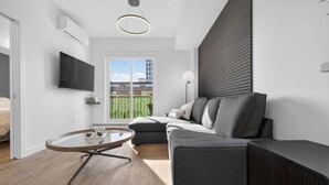 A modern living room with a grey sofa, a glass coffee table, a flat-screen TV, and a large window with a view of buildings. - 2BR | DT | Balcony | AC | Mins to Saddledome (Calgary)