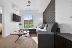 A modern living room with a grey sofa, a glass coffee table, a flat-screen TV, and a large window with a view of buildings. - 2BR | DT | Balcony | AC | Mins to Saddledome (Calgary)