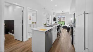 A modern kitchen with white cabinetry, a central island, and a dining area with a black sofa. - 2BR | DT | Balcony | AC | Mins to Saddledome (Calgary)