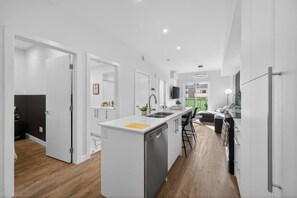 A modern kitchen with white cabinetry, a central island, and a dining area with a black sofa. - 2BR | DT | Balcony | AC | Mins to Saddledome (Calgary)