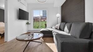 A modern living room with a grey sofa, a round glass coffee table, a flat-screen TV mounted on the wall, and a window with a view of a building and greenery. - 2BR | DT | Balcony | AC | Mins to Saddledome (Calgary)