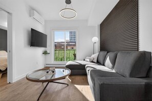 A modern living room with a grey sofa, a round glass coffee table, a flat-screen TV mounted on the wall, and a window with a view of a building and greenery. - 2BR | DT | Balcony | AC | Mins to Saddledome (Calgary)