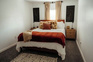 Un dormitorio con cama, dos mesitas de noche, una mesita auxiliar, una ventana con cortinas y obras de arte en las paredes.