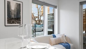 Uma área de refeições com uma mesa posta para dois, um sofá com almofadas, uma janela com persianas e um quadro na parede.