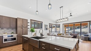 A modern kitchen with wooden cabinets, a large island, and a view of a snowy landscape through large windows. - Summit County Gem |  Designer Kitchen + Great Views (Silverthorne)