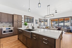 A modern kitchen with wooden cabinets, a large island, and a view of a snowy landscape through large windows. - Summit County Gem |  Designer Kitchen + Great Views (Silverthorne)