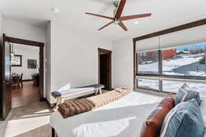 A bedroom with a large bed, a ceiling fan, a bench, and a view of a snowy landscape. - Summit County Gem |  Designer Kitchen + Great Views (Silverthorne)