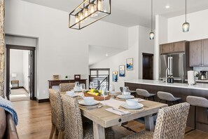 A modern dining area with a wooden table set for four, surrounded by patterned chairs, a kitchen with stainless steel appliances, and a hanging light fixture. - Summit County Gem |  Designer Kitchen + Great Views (Silverthorne)
