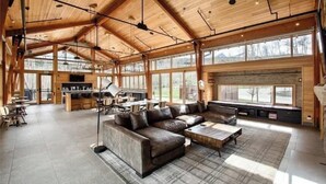 A spacious living area with a large leather sofa, a coffee table, and a dining table. There are large windows providing natural light and views of the outdoors. - Summit County Gem |  Designer Kitchen + Great Views (Silverthorne)