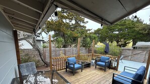 Una terraza cubierta al aire libre con suelo de madera, dos sillas azules con cojines, una mesa de vidrio y una valla de madera.