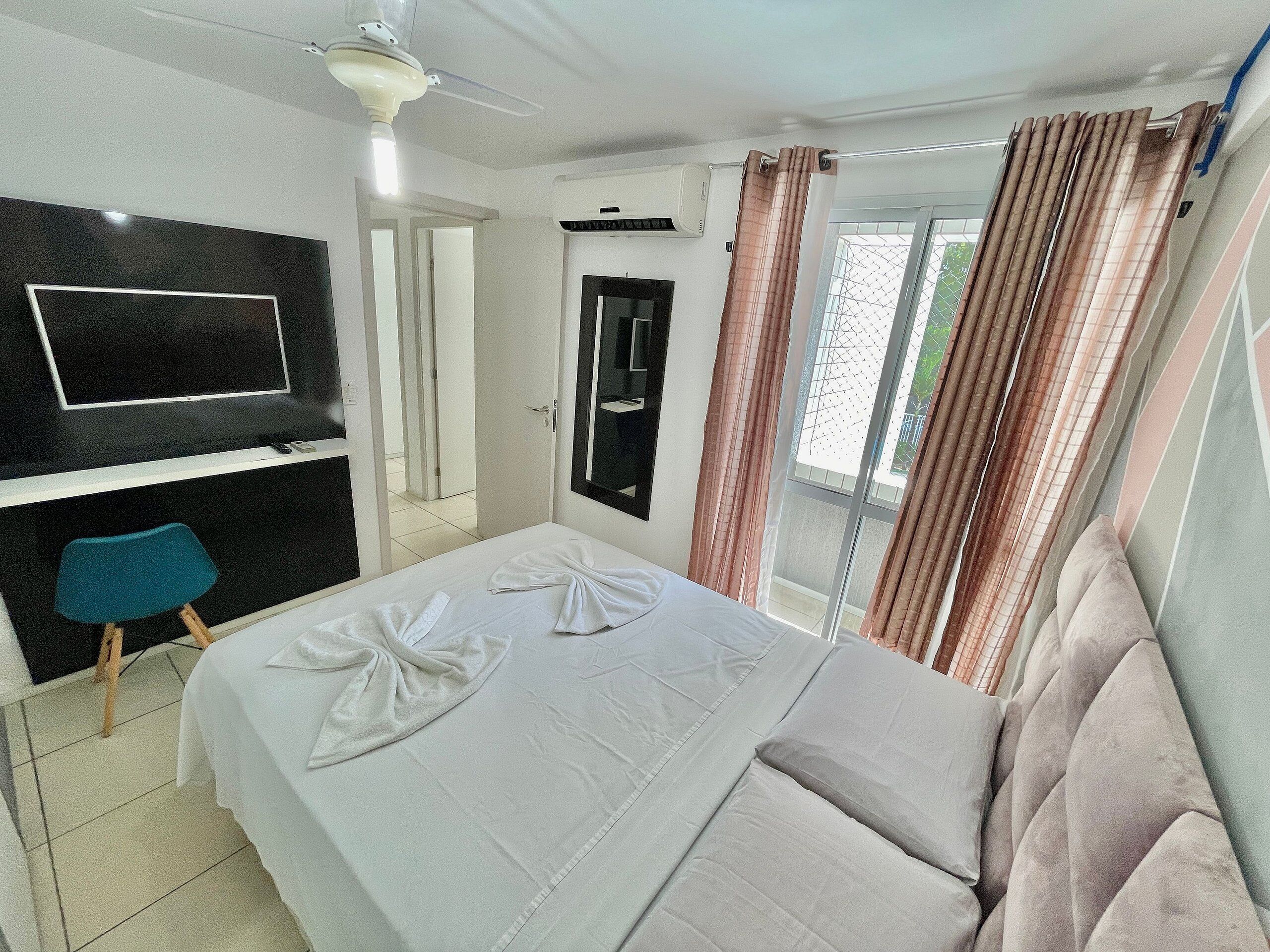 A hotel room with a bed, a TV mounted on the wall, a bathroom mirror, and a window with curtains.