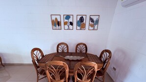 Une table de salle à manger ronde en bois, accompagnée de six chaises, un sol carrelé de carreaux blancs et quatre œuvres d’art représentant des feuilles, encadrées et accrochées au mur.