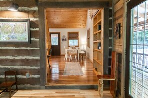 A log cabin interior with a wooden floor, a dining area with a table and chairs, and a bookshelf.