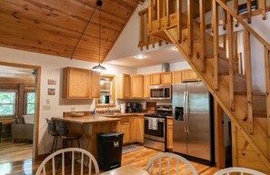Una cocina con armarios de madera, un refrigerador de acero inoxidable, una escalera de madera y un comedor con sillas.