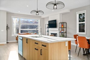 A modern kitchen with a large island, a dining area with orange chairs, and a TV mounted on the wall. - 16 Guests | 8BR | Game Rooms | King Beds (Calgary)