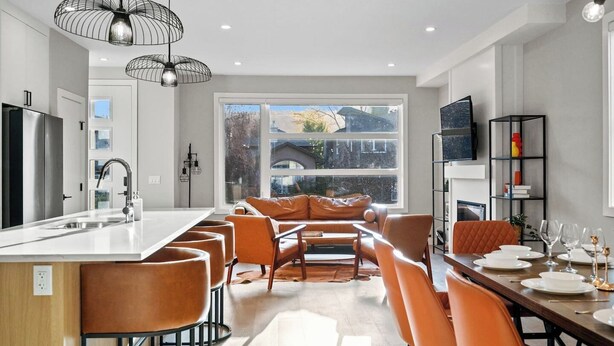 Een moderne keuken met een centraal eiland, een eethoek met oranje stoelen en een groot raam met uitzicht op bomen en een huis.