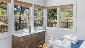 Un dormitorio con un aparador de madera, una cama con ropa de cama blanca y azul, y ventanas grandes con vistas a árboles y una valla.