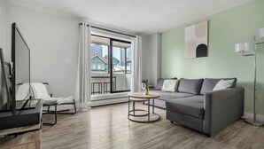 Een moderne woonkamer met een grijze bank, een klein rond tafeltje, een televisie en een groot raam met gordijnen.