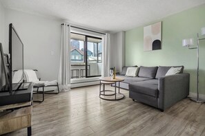 A modern living room with a grey sofa, a small round table, a TV, and a large window with curtains. - Min to Saddledome | 2 Bed | Central (Calgary)
