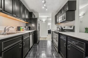 A modern kitchen with black cabinets, stainless steel appliances, and a tiled floor. - Min to Saddledome | 2 Bed | Central (Calgary)