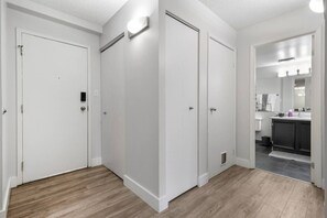 A clean, modern apartment with white walls, a wooden floor, and a kitchen area visible through an open door. - Min to Saddledome | 2 Bed | Central (Calgary)