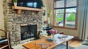 A stone fireplace with a flat-screen TV above, a wooden table with books and a vase of flowers, and a window with blinds. - Mountain Retreat: Privacy & Comfort at 4,400 ft (Mars Hill)