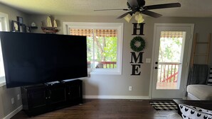 Een woonkamer met een flatscreen televisie op een zwarte televisiemeubel, een plafondventilator, en een 'HOME' bord aan de muur.