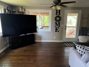 Un salon avec un téléviseur à écran plat posé sur un meuble noir, un ventilateur de plafond et un panneau décoratif portant l’inscription « HOME » au mur.